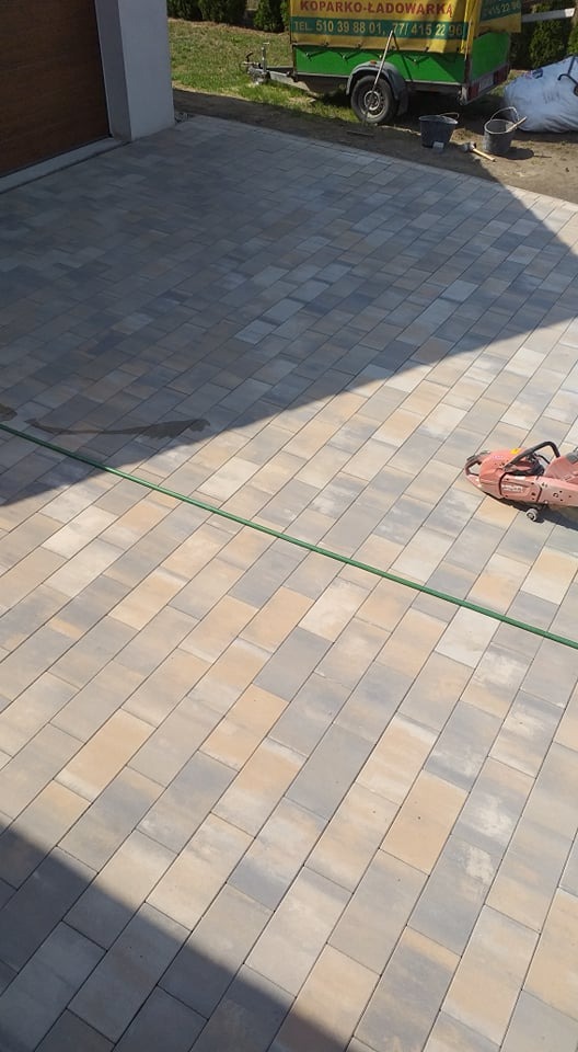 Ułożona kostka brukowa w odcieniach szarości i beżu, z widocznym wibratorem do ubijania i zielonym wężem ogrodowym, przy garażu i przyczepie z napisem.