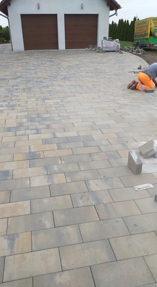 Ułożona kostka brukowa w odcieniach szarości i beżu na podjeździe przed garażem, widoczny pracownik w pomarańczowych spodniach i ciężarówka firmy w tle.