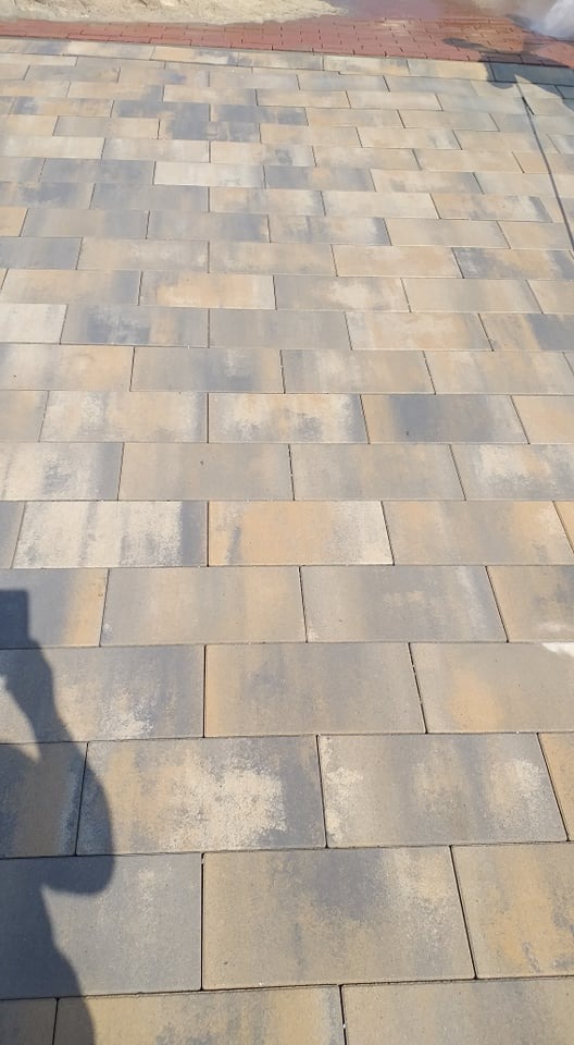 Ułożona kostka brukowa w odcieniach szarości i beżu, z widocznym cieniem osoby robiącej zdjęcie, na obrzeżu widoczne czerwone cegły.