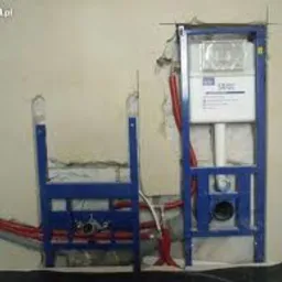 Stelaż podtynkowy do WC w trakcie montażu, widoczne niepodłączone rury hydrauliczne i fragment ściany z uszkodzonym tynkiem.