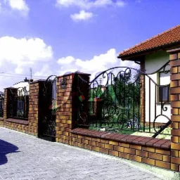 Ozdobna brama wjazdowa z kutego żelaza, osadzona w murze z cegły klinkierowej, Warszawa. Widoczny fragment chodnika z kostki brukowej i fragment domu w tle.