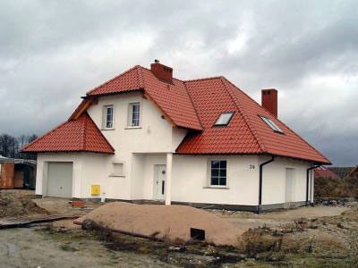 Nowo wybudowany, biały dom jednorodzinny z czerwoną dachówką, widoczny z zewnątrz, otoczony piaskiem i gruzem budowlanym, na tle pochmurnego nieba.