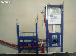 Stelaż podtynkowy do WC w trakcie montażu, widoczne niepodłączone rury hydrauliczne i fragment ściany z uszkodzonym tynkiem.