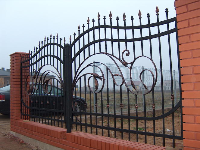 Kuta brama ogrodzeniowa z dekoracyjnymi elementami w kształcie liści i grotów, osadzona na ceglanych słupkach, widok z zewnątrz posesji.