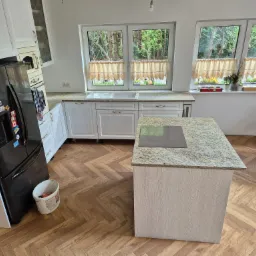 Kuchnia z wyspą i blatem o wzorze kamienia, parkiet ułożony w jodełkę, widok na ogród przez okno.