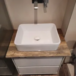 Nowoczesna łazienka z kwadratową białą umywalką nablatową, chromowaną armaturą i blatem imitującym trawertyn, zamontowaną na szafce z szufladami.