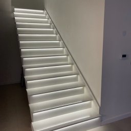 FUPH-MGP - Nowoczesne, białe schody z podświetleniem LED wzdłuż stopni, oświetlające minimalistyczne wnętrze z jasnymi ścianami i podłogą.