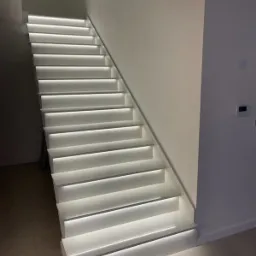 Nowoczesne, białe schody z podświetleniem LED wzdłuż stopni, oświetlające minimalistyczne wnętrze z jasnymi ścianami i podłogą.