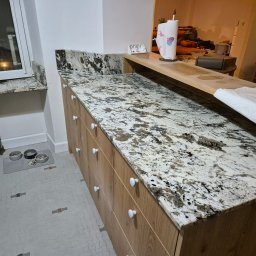 FUPH-MGP - Kuchnia z blatem z jasnego granitu z ciemnymi żyłkami, drewniane szafki z białymi gałkami, w tle widoczny fragment salonu i miski dla zwierząt przy grzejniku.