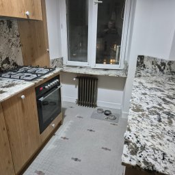 FUPH-MGP - Kuchnia z drewnianymi szafkami, blatem i okładziną ściany z jasnego kamienia z ciemnymi wzorami, okno z parapetem z tego samego kamienia i miski dla zwierząt na podłodze.