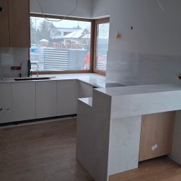 FUPH-MGP - Jasna kuchnia z blatem imitującym marmur, widok na okno z zabudową. Elementy wykończeniowe w trakcie montażu. Na zewnątrz widać budowę.