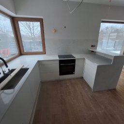 FUPH-MGP - Jasna kuchnia z blatem z białego kamienia, czarną armaturą i drewnianą podłogą. Widoczne okna z drewnianymi ramami i fragment placu budowy za nimi. Minimalistyczny design.