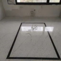 Wnętrze łazienki w trakcie remontu, z jasnymi płytkami podłogowymi z ciemną bordiurą tworzącą ramę, widoczne kable elektryczne na podłodze, surowy stan ścian z widocznym oknem.