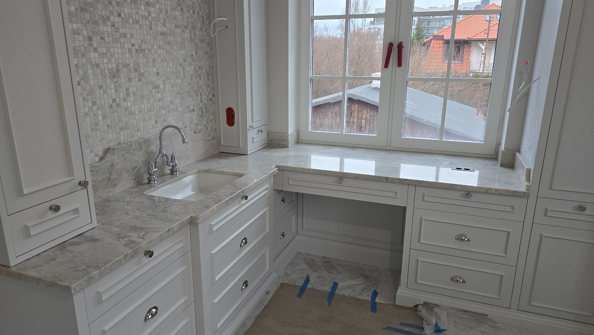 Jasny blat z kamienia naturalnego z wbudowanym zlewem i miejscem do pracy przy oknie, białe szafki z frezowanymi frontami i chromowanymi uchwytami, mozaika na ścianie, widok na zabudowania za oknem.