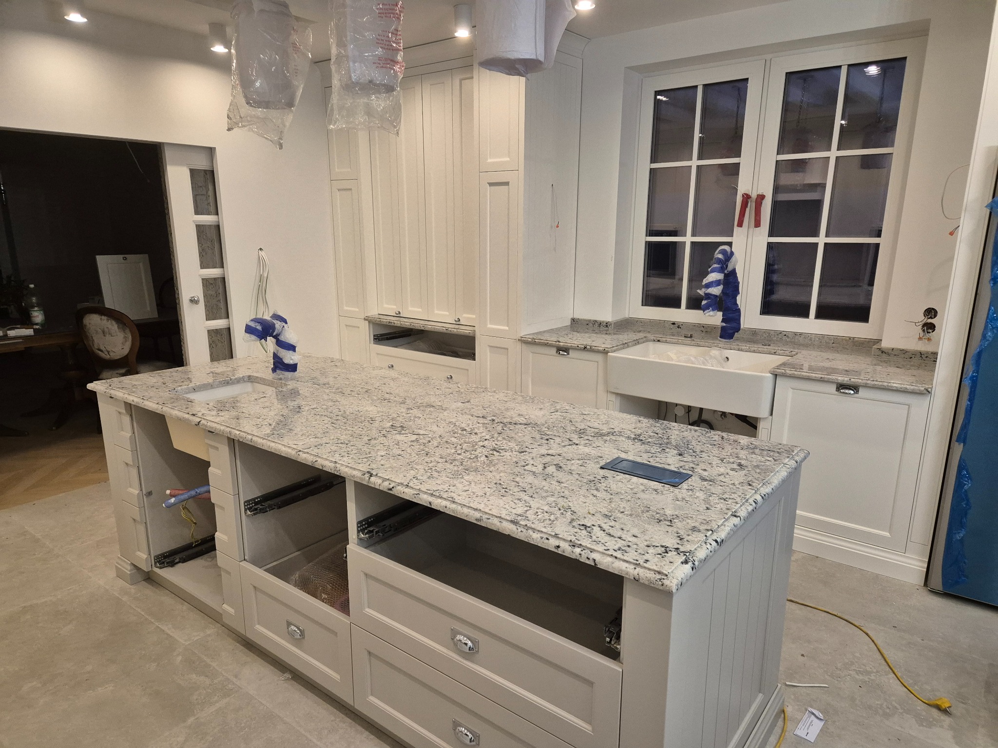 Wnętrze kuchni w trakcie montażu, z wyspą z jasnym, nakrapianym blatem i białymi szafkami. Otwarta szuflada wyspy ukazuje zabezpieczone elementy, a okno nad zlewem jest częściowo zasłonięte folią.