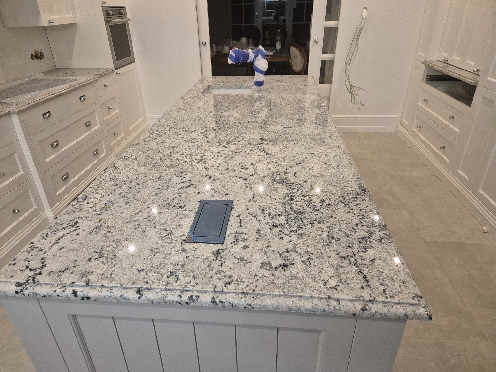 Nowa, jasna kuchnia z wyspą i blatem o wzorze soli i pieprzu, bateria zaklejona taśmą, widoczne gniazdo elektryczne w blacie