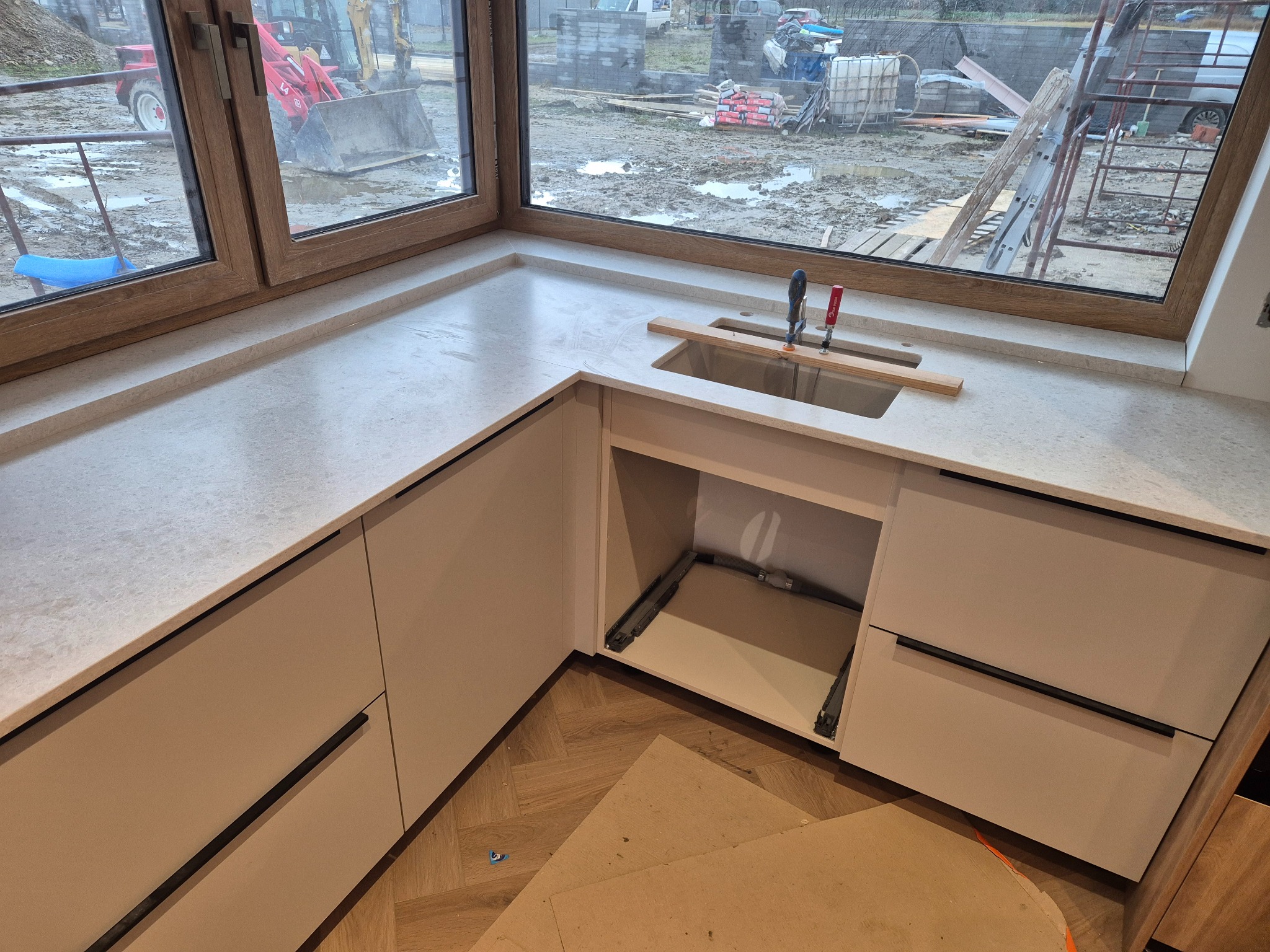 Jasny, narożny blat w kuchni, zamontowany pod oknem z widokiem na plac budowy, z widocznym zlewem i narzędziami montażowymi.