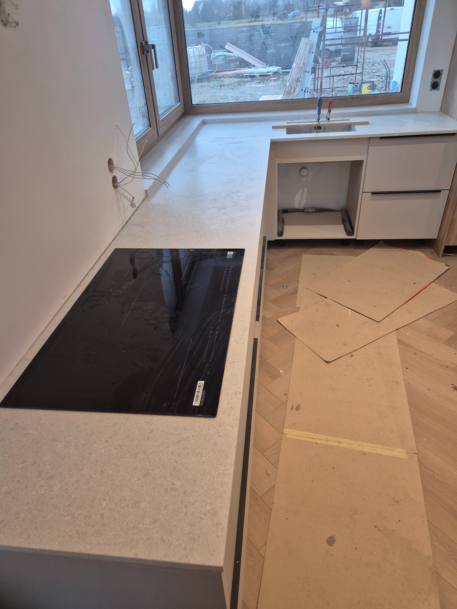 Jasny blat kamienny w kuchni z zamontowaną płytą indukcyjną, widoczne przygotowania do montażu zlewu i zabezpieczenia podłogi kartonem.