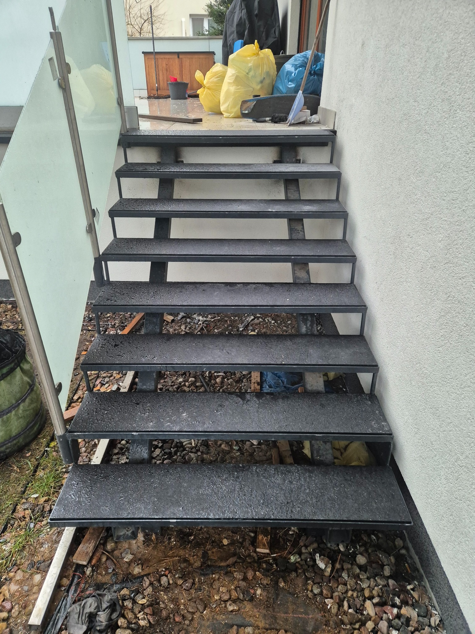 Metalowe schody zewnętrzne z szarym wykończeniem, widoczne ślady po deszczu, częściowo osłonięte szklaną balustradą, teren wokół w trakcie prac wykończeniowych.