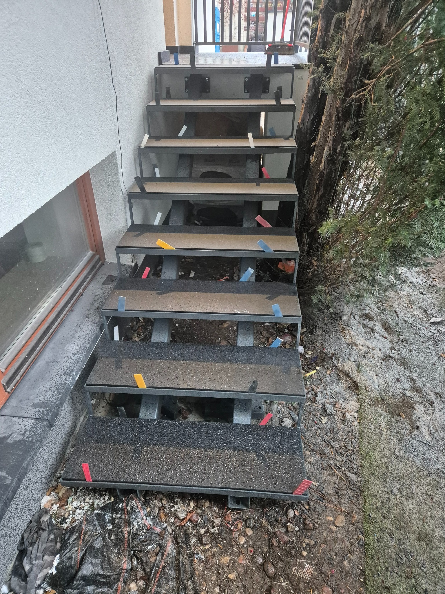Konstrukcja schodów zewnętrznych z metalową ramą i tymczasowymi elementami mocującymi, widoczne krople wody na powierzchni stopni, w tle ściana budynku i zieleń.