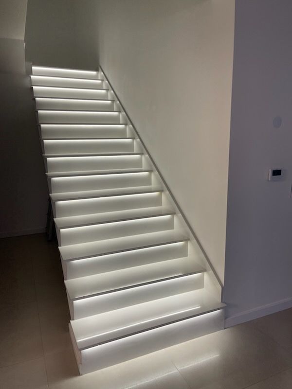 Nowoczesne, białe schody z podświetleniem LED wzdłuż stopni, oświetlające minimalistyczne wnętrze z jasnymi ścianami i podłogą.