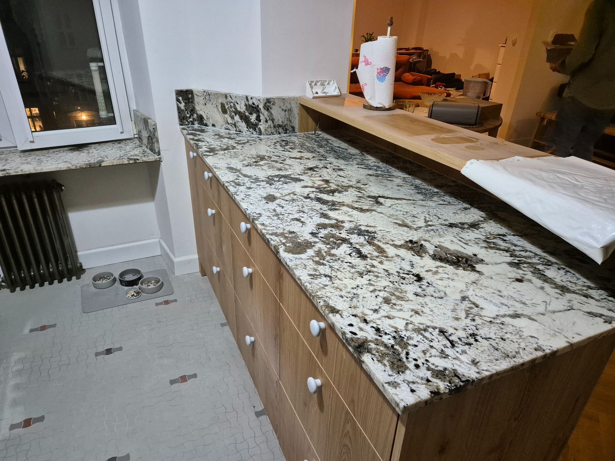 Kuchnia z blatem z jasnego granitu z ciemnymi żyłkami, drewniane szafki z białymi gałkami, w tle widoczny fragment salonu i miski dla zwierząt przy grzejniku.