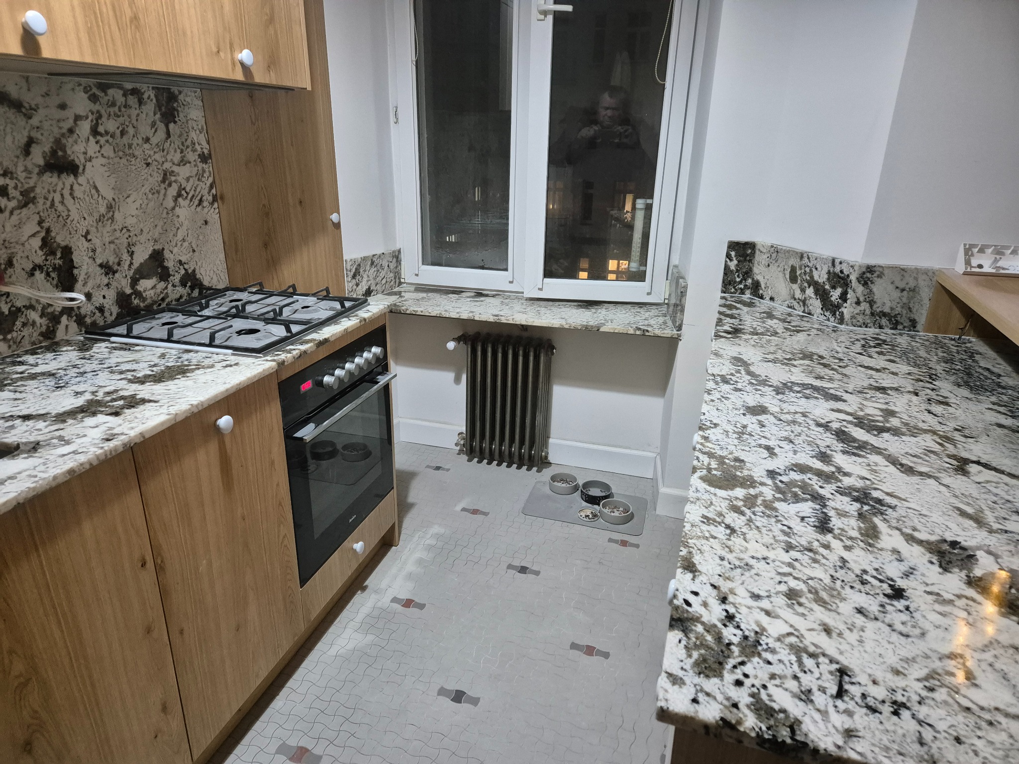 Kuchnia z drewnianymi szafkami, blatem i okładziną ściany z jasnego kamienia z ciemnymi wzorami, okno z parapetem z tego samego kamienia i miski dla zwierząt na podłodze.