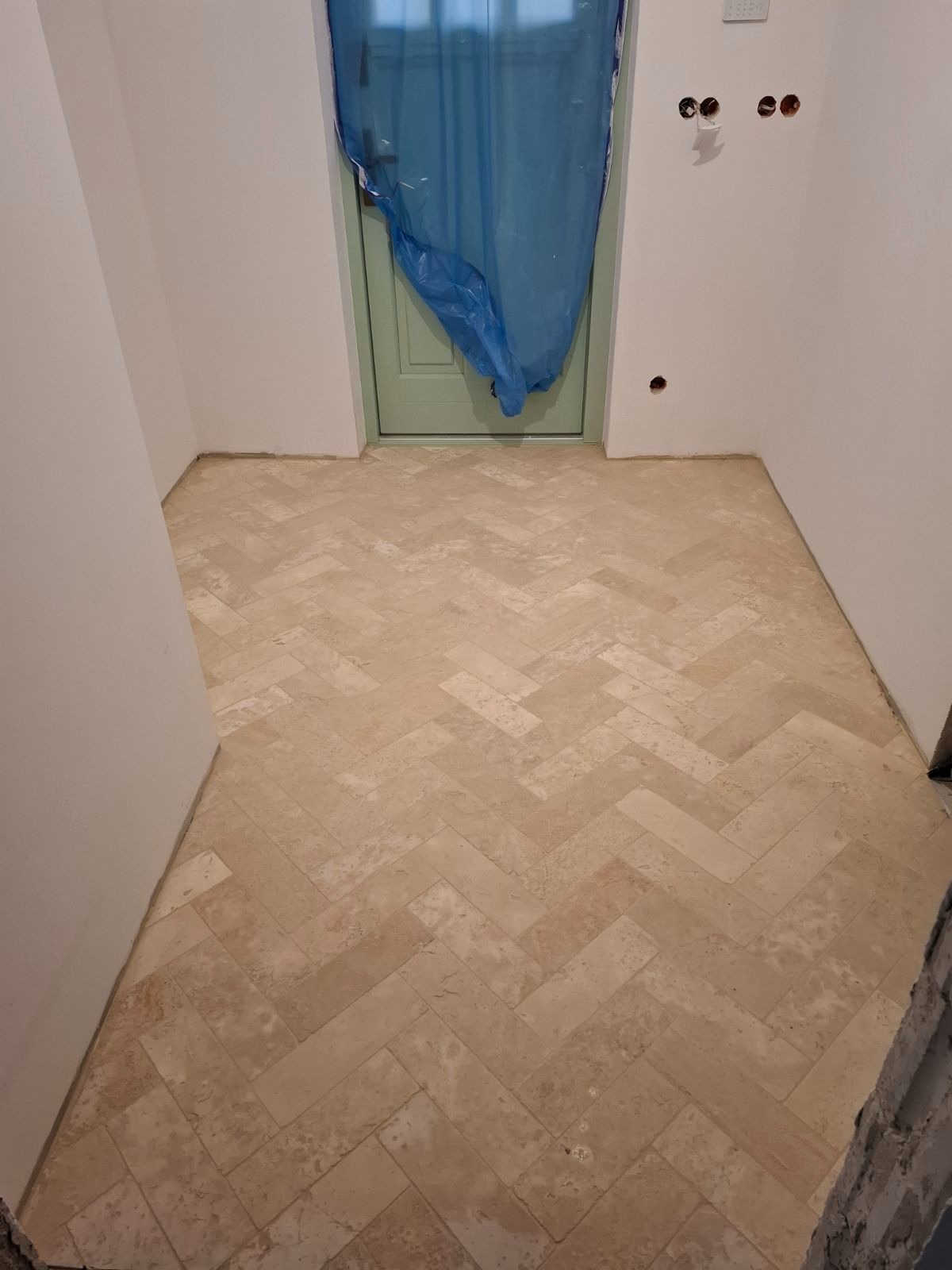 Ułożona podłoga z beżowych płytek w jodełkę, widoczna niebieska folia zabezpieczająca drzwi, ściany w trakcie wykańczania.