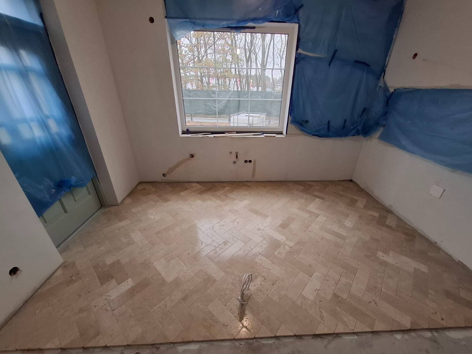 Ułożona podłoga z kamiennych płytek w jodełkę w jasnym odcieniu, ściany w trakcie przygotowania do malowania, zabezpieczone folią, widoczne wystające kable i rury instalacyjne.