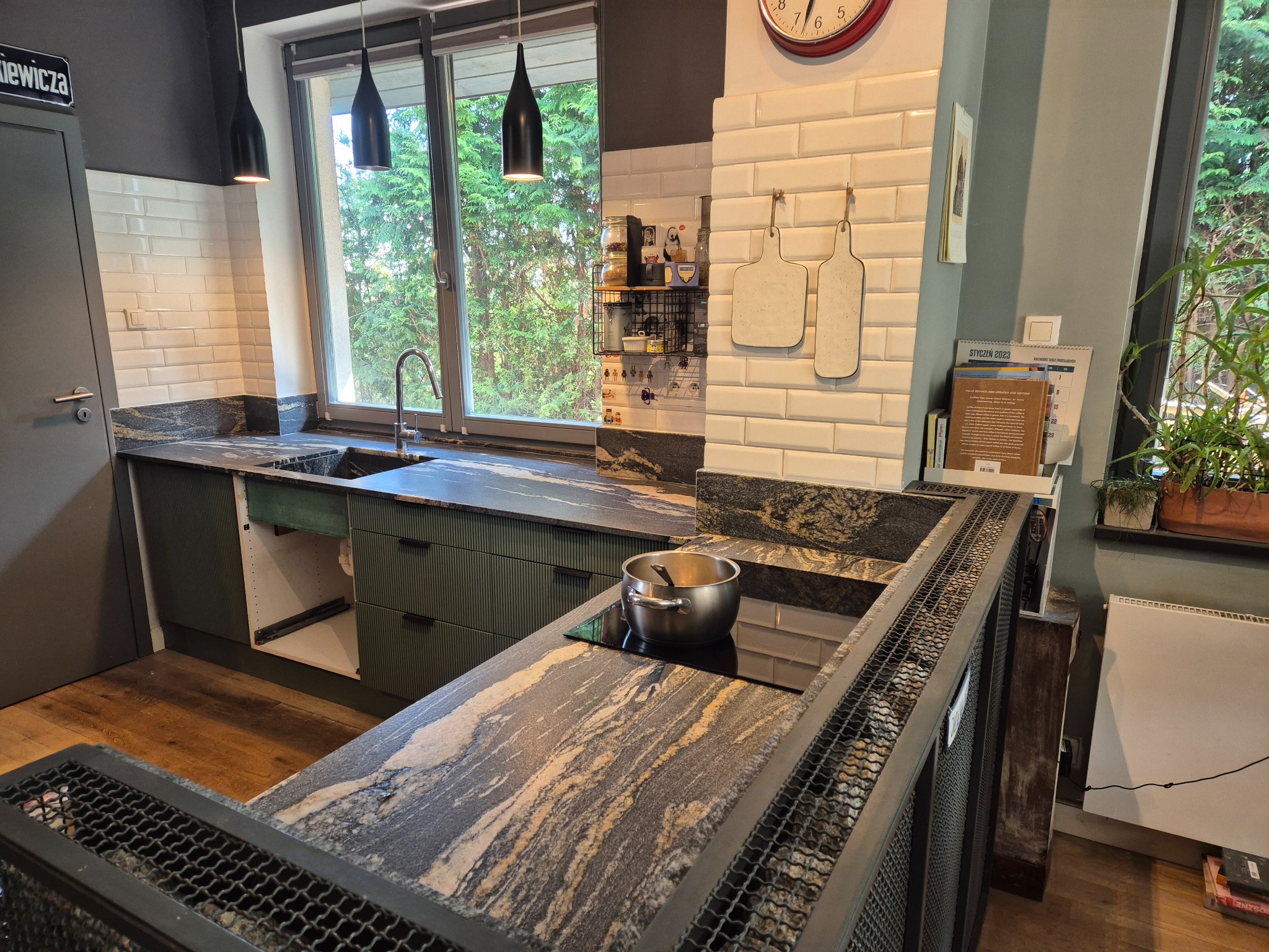 Kuchenny blat z granitu o unikalnym wzorze, z widocznym garnkiem na płycie indukcyjnej, nowoczesne, wiszące lampy i zielone szafki w tle.