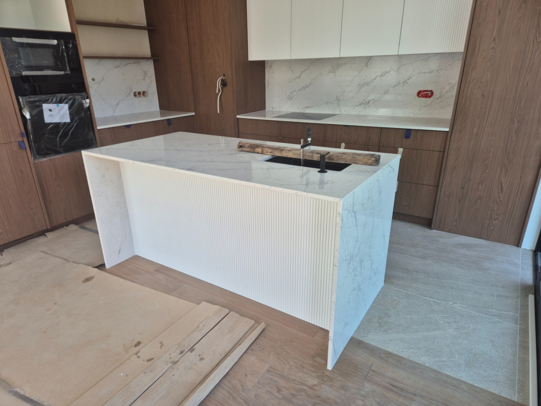 Wnętrze kuchni w Skarżysko-Kamiennej w trakcie montażu, z marmurowym blatem wyspy kuchennej o ryflowanym wykończeniu, zabezpieczonym drewnianymi belkami wokół wbudowanego zlewu, na podłodze leżą...