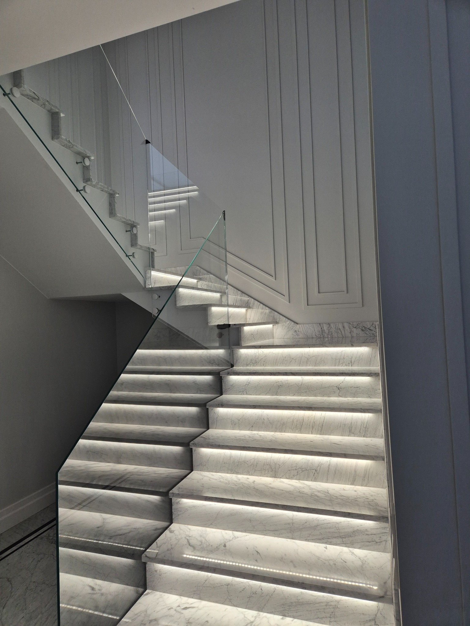 Nowoczesne schody kamienne z podświetleniem LED i szklaną balustradą, marmur Carrara, minimalistyczny design, białe ściany z dekoracyjnymi listwami.