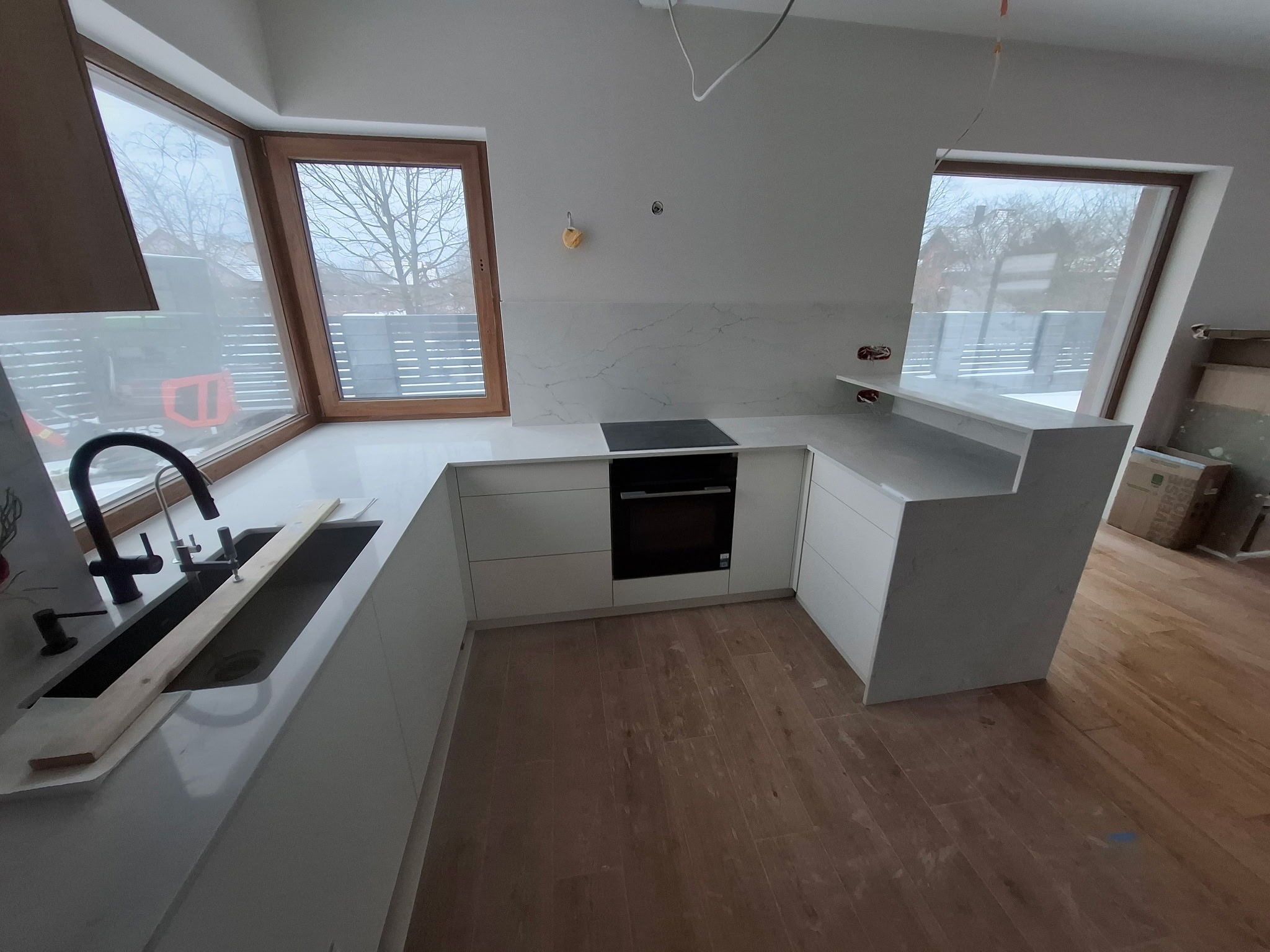 Jasna kuchnia z blatem z białego kamienia, czarną armaturą i drewnianą podłogą. Widoczne okna z drewnianymi ramami i fragment placu budowy za nimi. Minimalistyczny design.