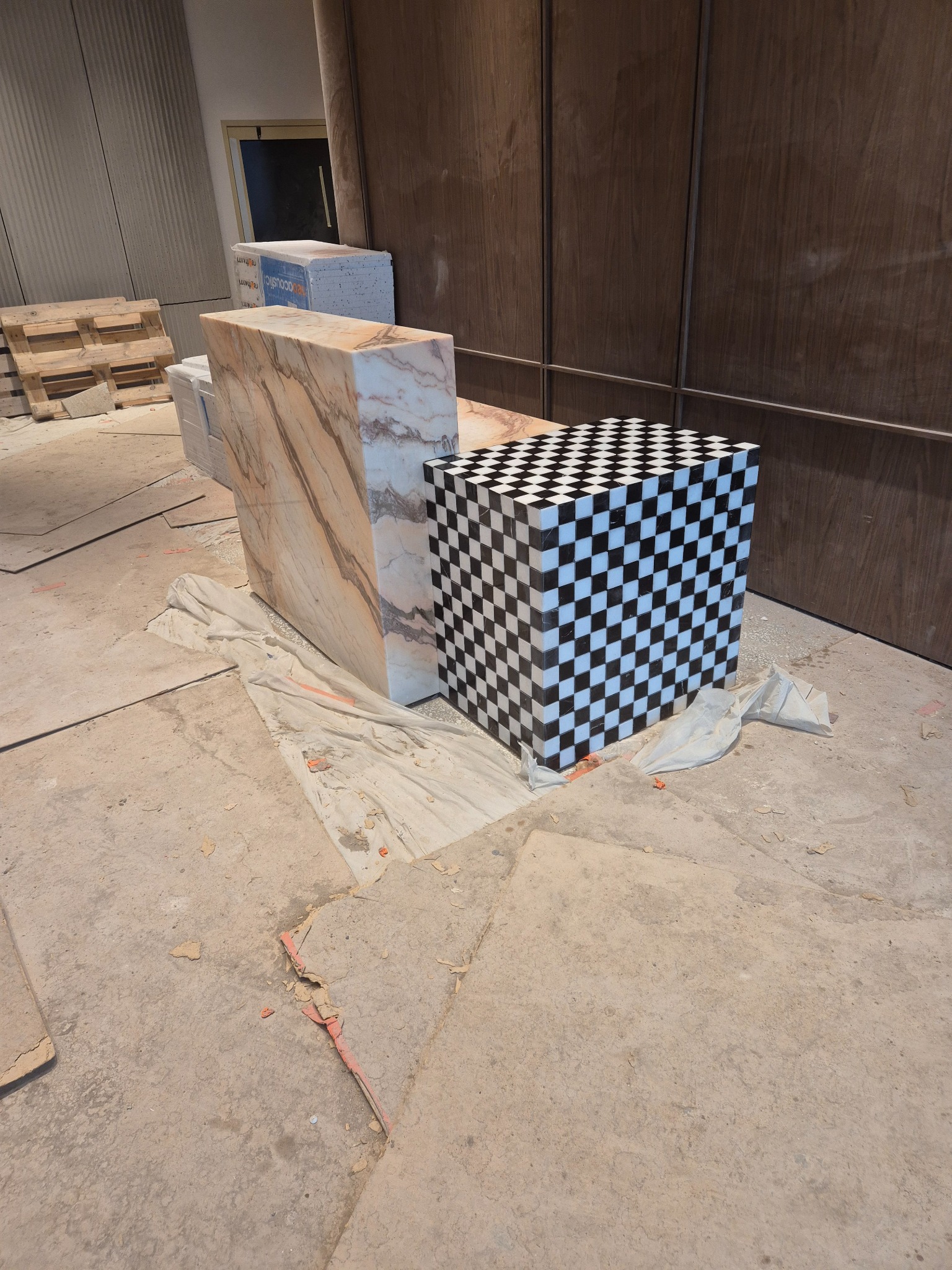 Bloki z kamienia: marmur w odcieniach beżu i szachownica czarno-biała, ustawione na surowym betonie w pomieszczeniu w trakcie remontu. Widoczne palety i zabezpieczenia.