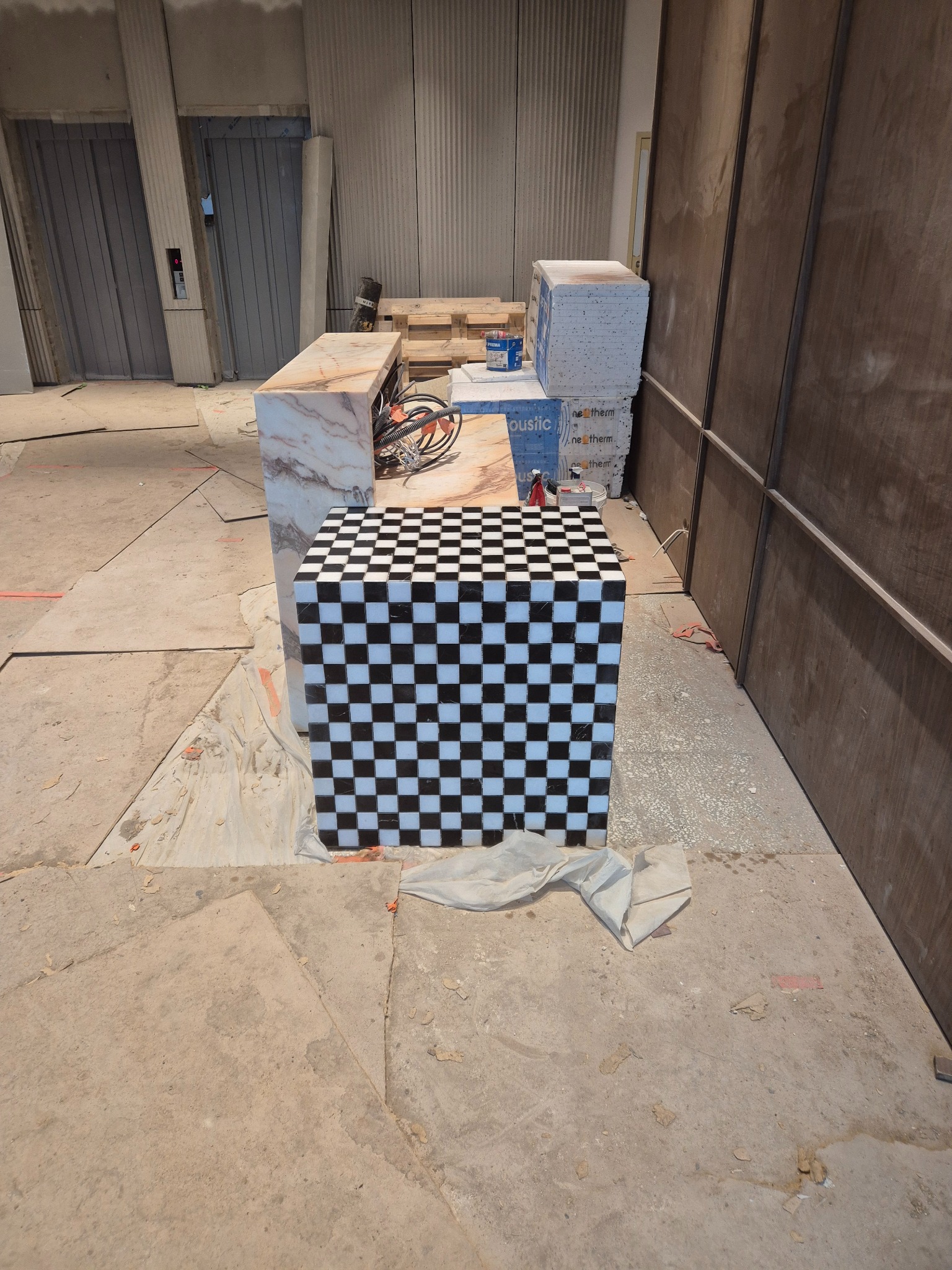 Niedokończony blat z marmuru i postument z czarno-białą szachownicą na budowie. Widoczne materiały budowlane i surowy beton. W tle drzwi windy.