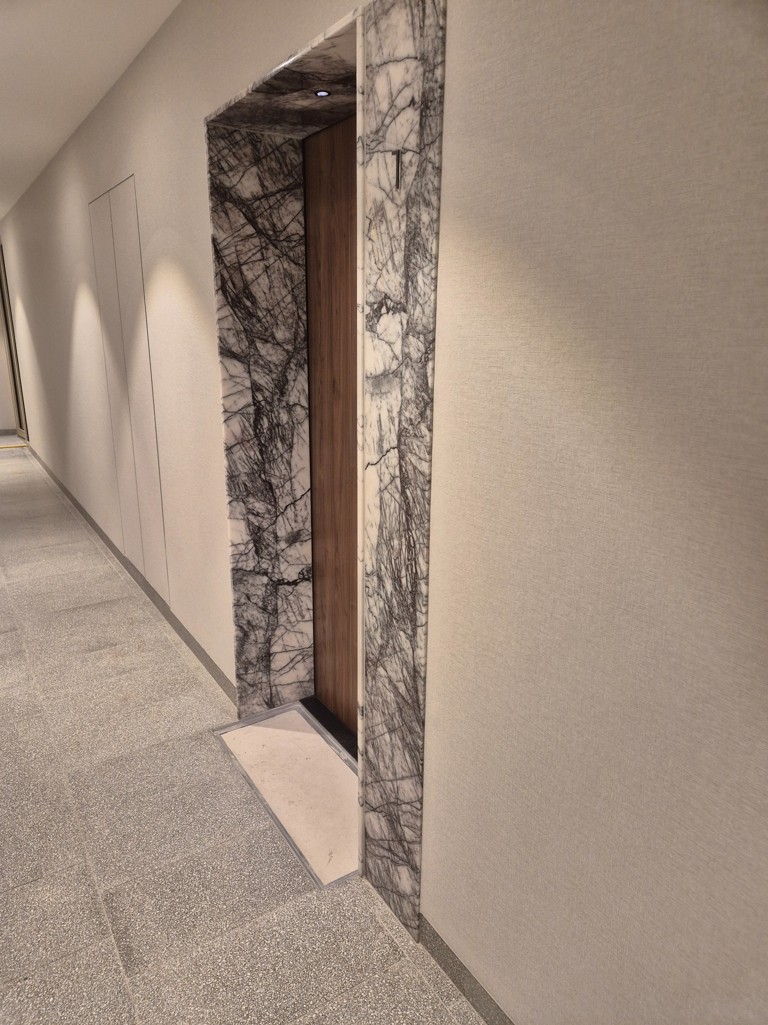 Wykończenie wejścia do windy marmurem o unikalnym wzorze żyłkowania. Eleganckie połączenie kamienia i drewna w nowoczesnym wnętrzu. Subtelne oświetlenie podkreśla detale.
