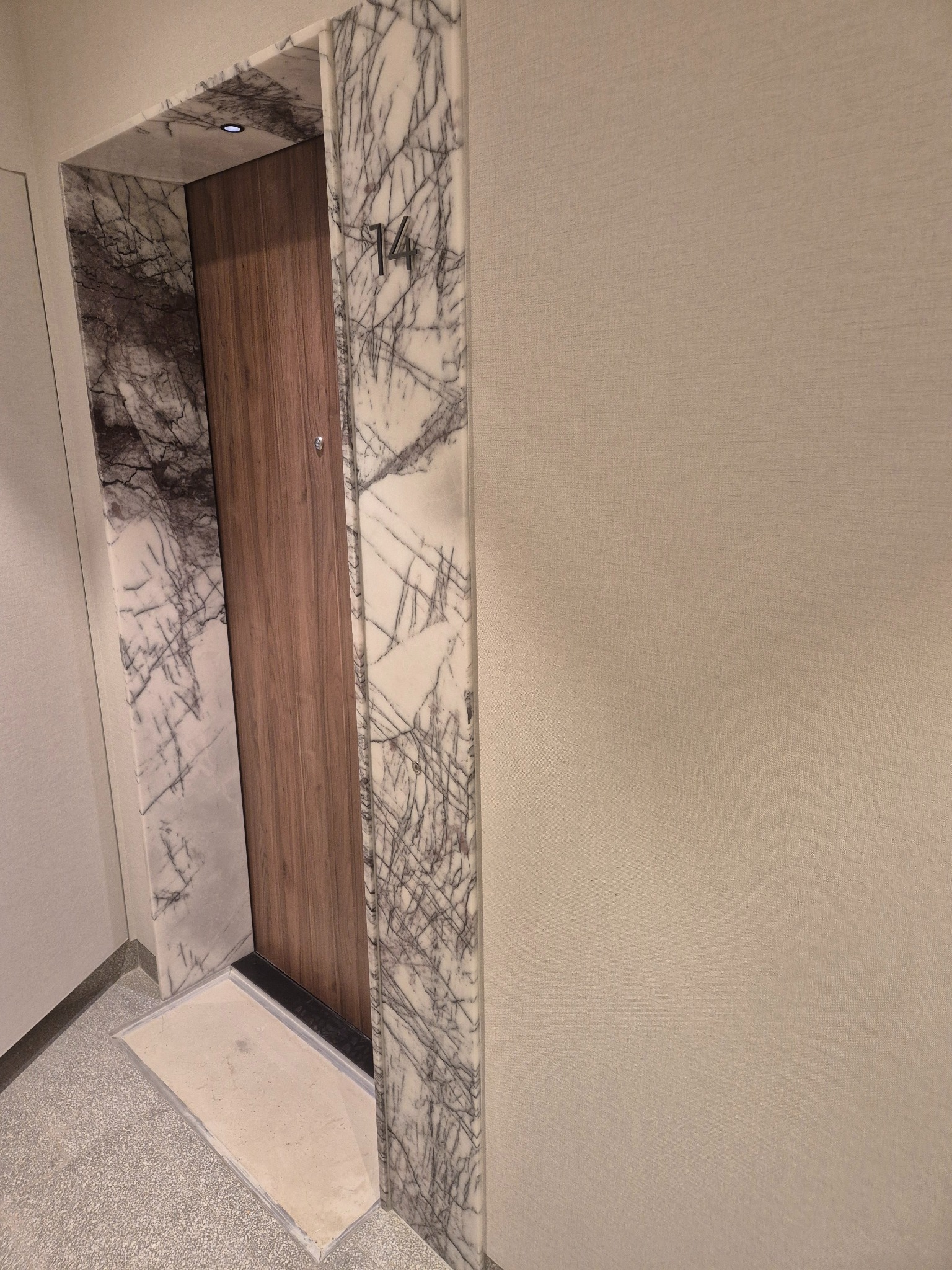 Wejście do mieszkania z drewnianymi drzwiami i marmurowym portalem z numerem '14'. Widoczne wykończenie podłogi i ściany w neutralnych barwach.