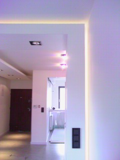 Nowoczesne wnętrze z białymi ścianami, oświetleniem LED wzdłuż sufitu i ścian, oraz ciemnymi elementami wykończenia, takimi jak drzwi i gniazdka.