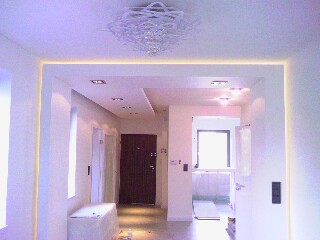 Nowoczesne wnętrze z białymi ścianami, oświetleniem LED wzdłuż sufitu i ozdobnym żyrandolem. Widoczny fragment korytarza z drzwiami wejściowymi i skrzynią owiniętą folią ochronną.