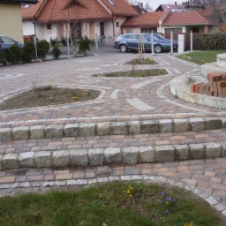 Ułożona kostka brukowa z elementami granitowymi tworząca schody i ścieżki z wyznaczonymi miejscami na zieleń, w tle domy i samochody.