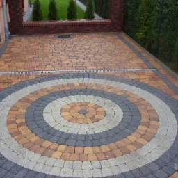 Kolorowa nawierzchnia z kostki brukowej ułożona w koncentryczne okręgi, z odpływem liniowym, obrzeżem i zielonym żywopłotem w tle.