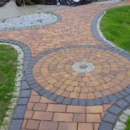 Ogród z ścieżką z brązowej kostki ułożonej w okrągły wzór, obramowaną szarą kostką, z elementami małej architektury i trawnikiem.