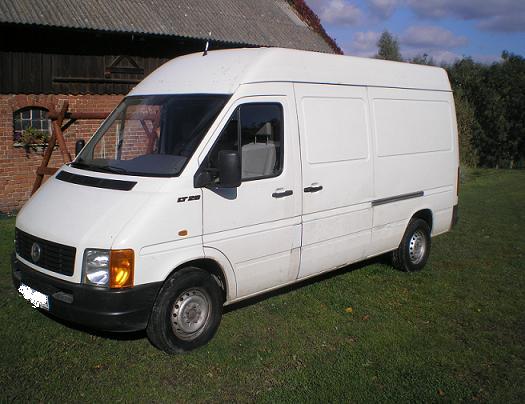 Biały bus LT 28 na trawie przed ceglanym budynkiem gospodarczym z drewnianymi elementami.