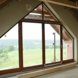 Poddasze z oknem o nietypowym trójkątnym kształcie, ściany po nałożeniu tynku, widoczne drewniane elementy konstrukcji dachu i krajobraz za oknem.