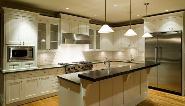 Jasna kuchnia na wymiar z białymi szafkami, granitowym blatem i wyspą kuchenną z ciemnym blatem, oświetlona trzema wiszącymi lampami stożkowymi.