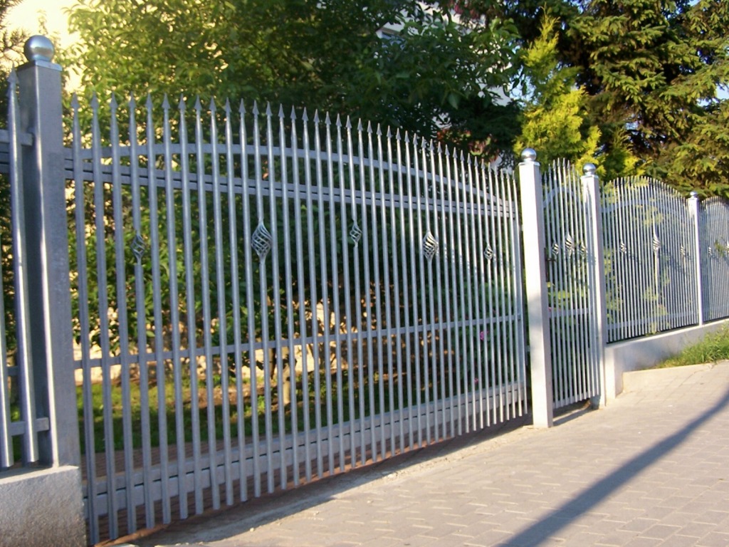 Metalowe ogrodzenie z pionowych prętów zakończonych szpicami, ozdobione skręconymi elementami dekoracyjnymi, widoczne z perspektywy chodnika.