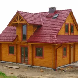 Gotowy, dwukondygnacyjny dom z drewna o żółtej elewacji i czerwonym dachu z dachówki, z widocznym kominem i oknem dachowym, na działce w trakcie zagospodarowania.