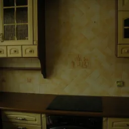 Kuchnia z kremowymi szafkami z przeszklonymi frontami i kwadratowymi, żółtymi płytkami na ścianie, ozdobionymi motywami butelek i kieliszków, nad blatem z wbudowaną płytą grzewczą.