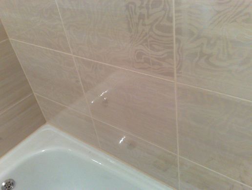 Fragment łazienki z beżowymi, wzorzystymi płytkami ceramicznymi na ścianie nad białą wanną z widocznym odpływem.