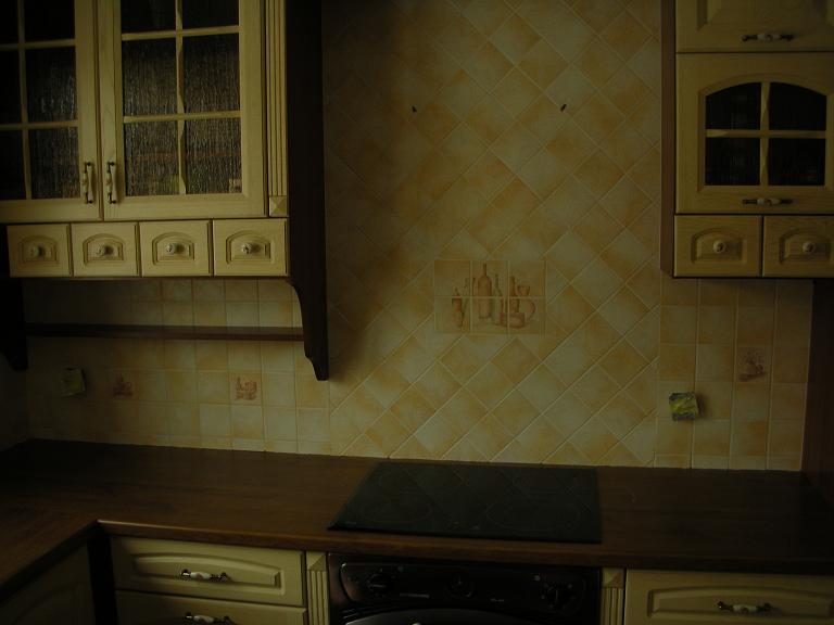 Kuchnia z kremowymi szafkami z przeszklonymi frontami i kwadratowymi, żółtymi płytkami na ścianie, ozdobionymi motywami butelek i kieliszków, nad blatem z wbudowaną płytą grzewczą.